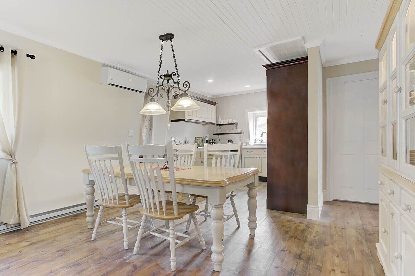 A dining area with a wooden table and chairs, a chandelier, and a kitchen in the background.