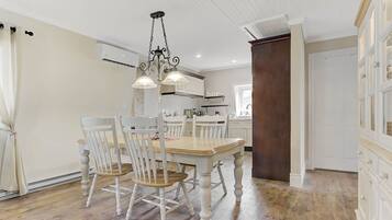 A dining area with a wooden table and chairs, a chandelier, and a kitchen in the background.