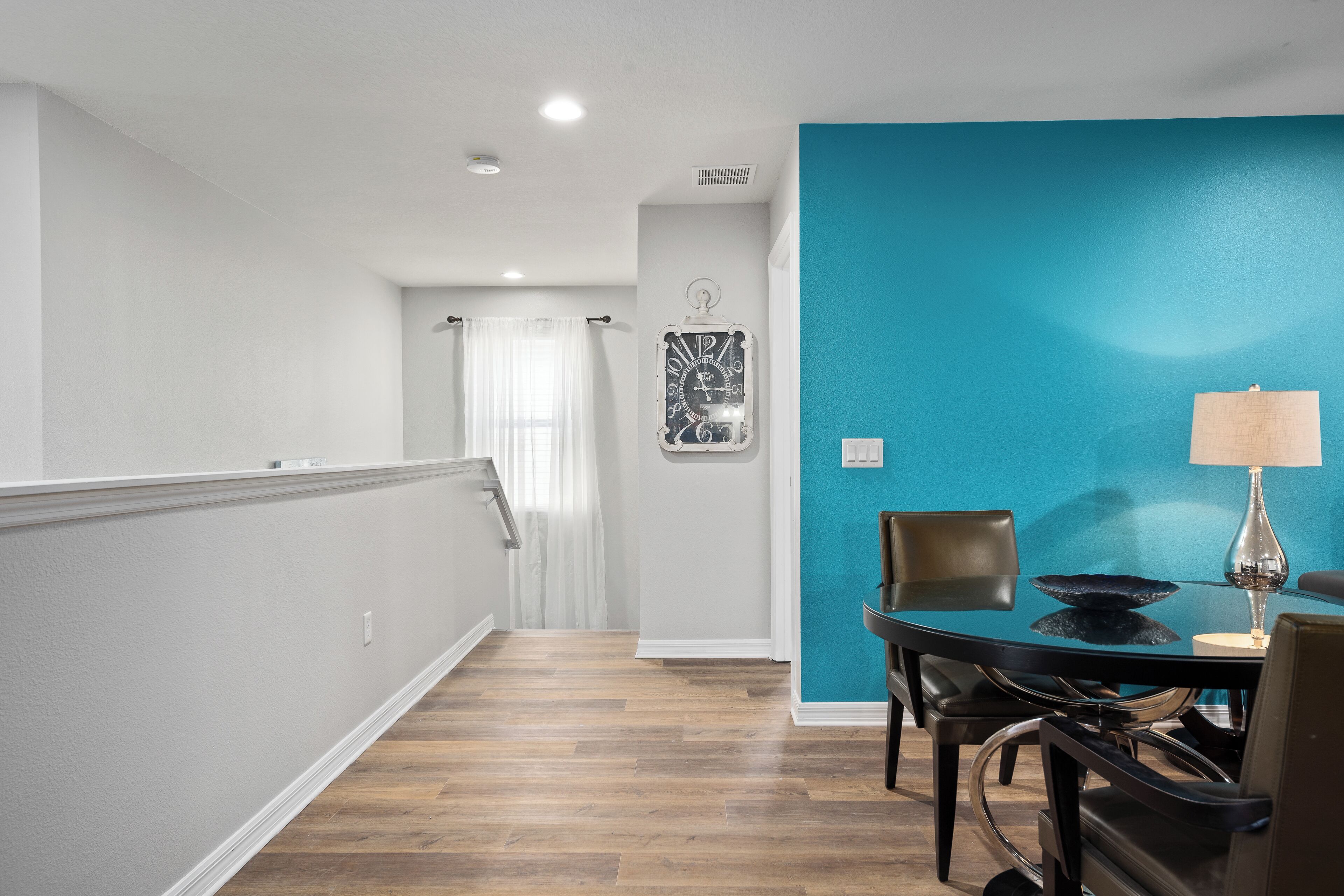 A modern interior with a wooden floor, a teal accent wall, a dining table with chairs, and a clock on the wall.