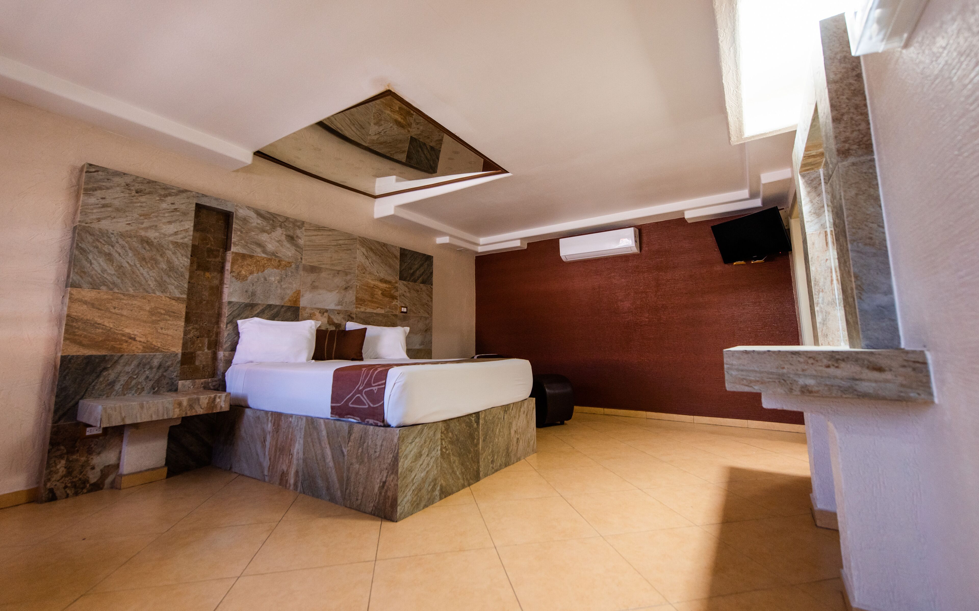 Habitación de hotel moderna con una pared de acento de piedra, una cama con ropa blanca, una mesita de noche pequeña y un banco empotrado.