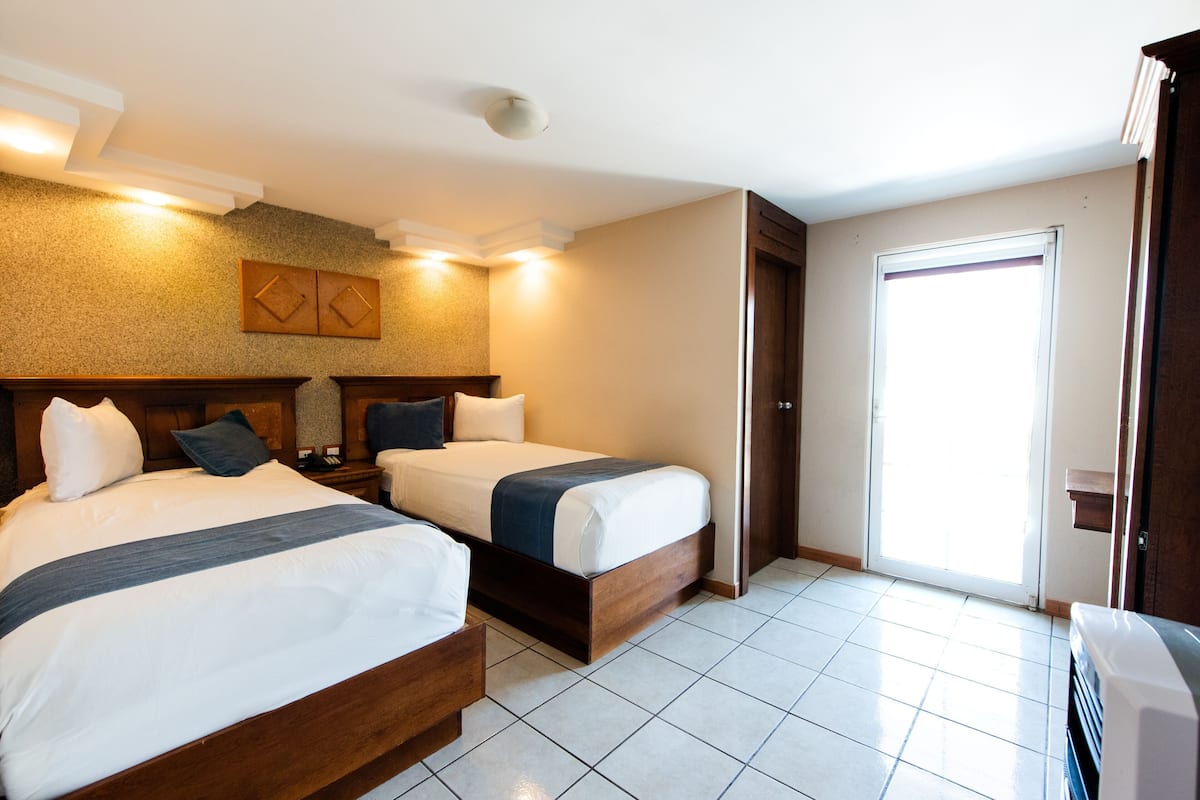 Habitación de hotel con dos camas, cabecero de madera, un ventanal grande y una puerta que da a un balcón.