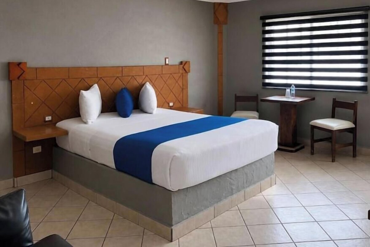 Una habitación de hotel con cama, mesitas de noche, una pequeña mesa con sillas y una ventana con persianas.