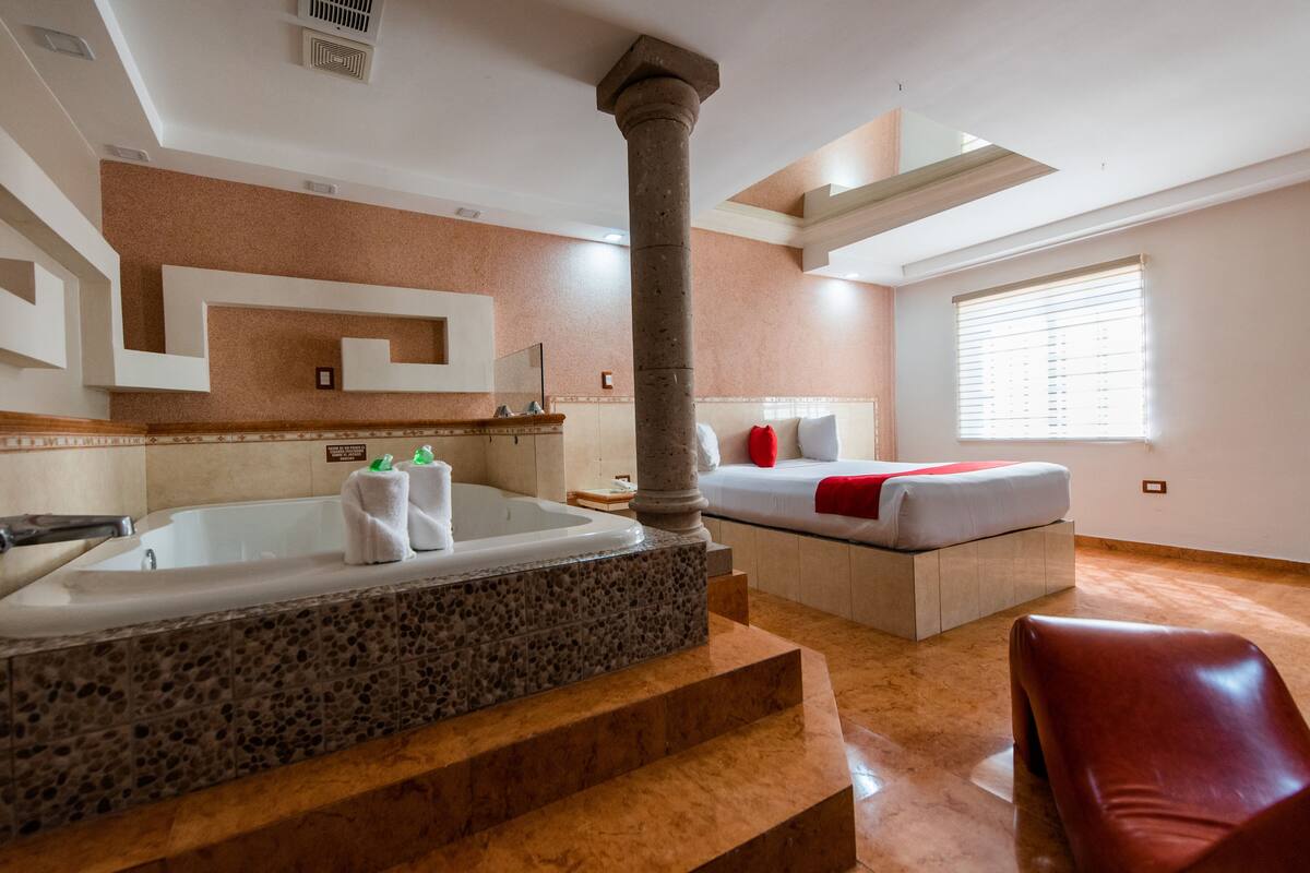 Habitación de hotel moderna con una bañera grande, una cama con una colcha roja y un suelo de mármol.