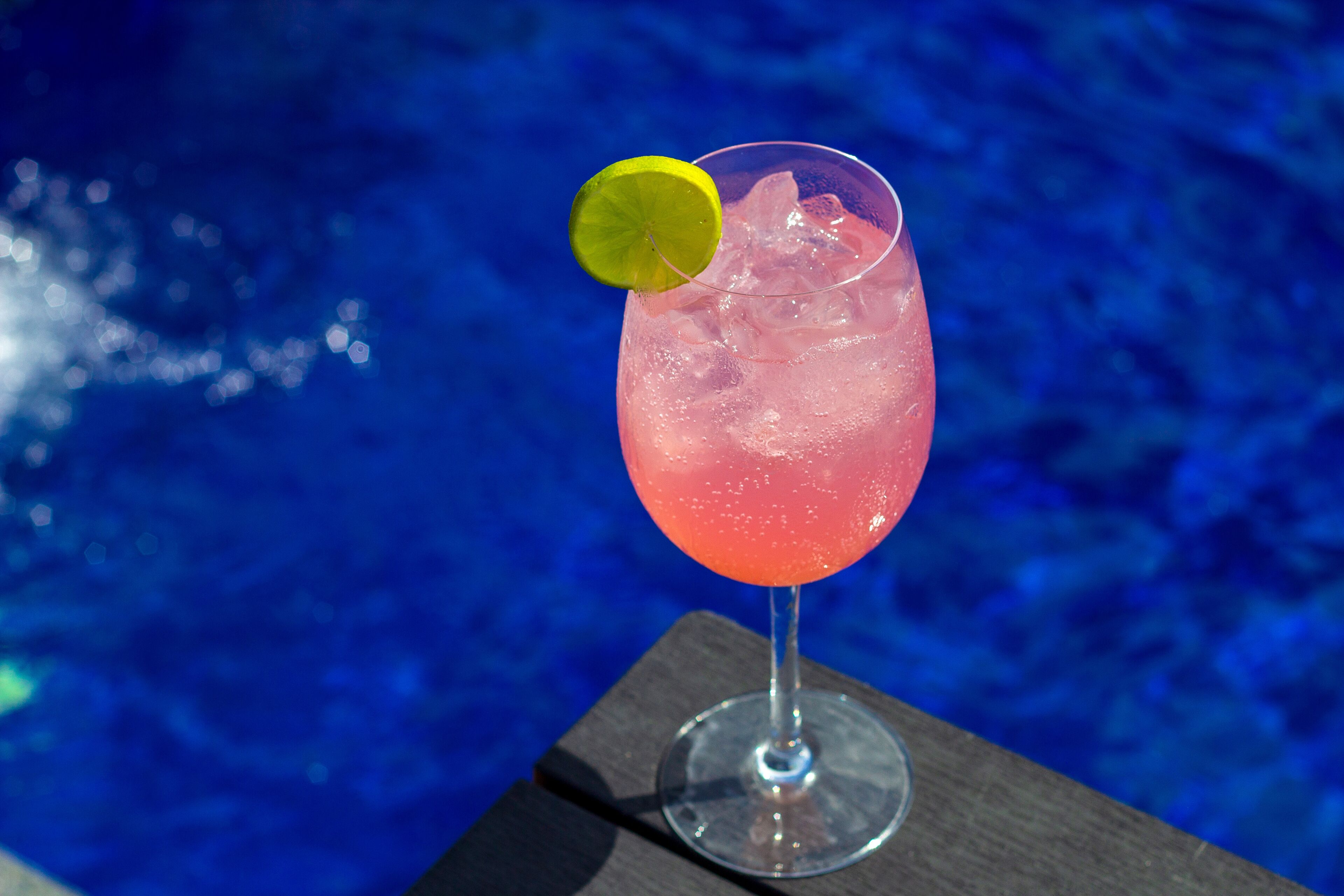 A pink cocktail with a lime garnish on a glass, placed on a wooden surface by a pool.