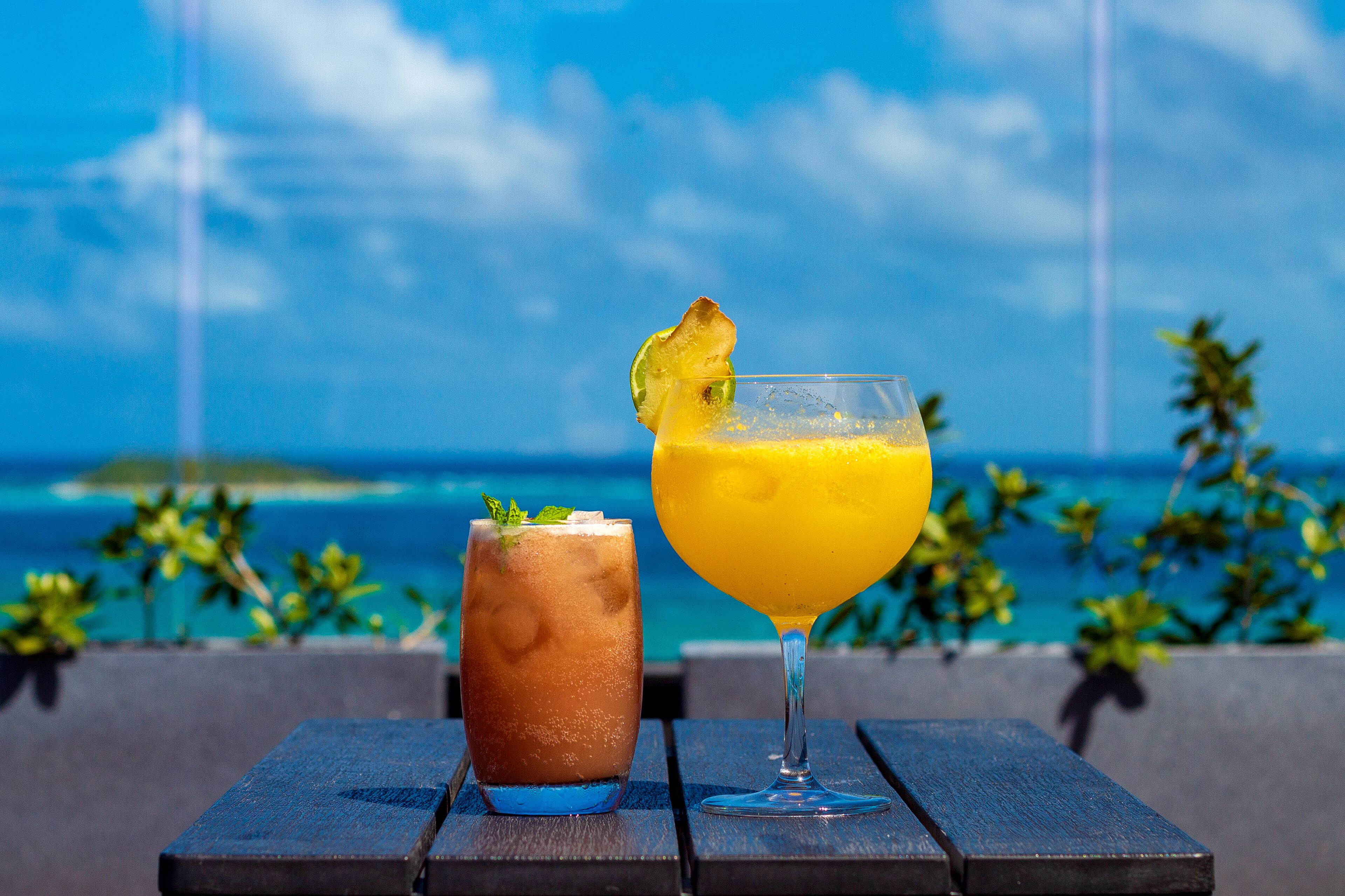 Dos bebidas sobre una mesa de madera con vista al océano y palmeras al fondo.