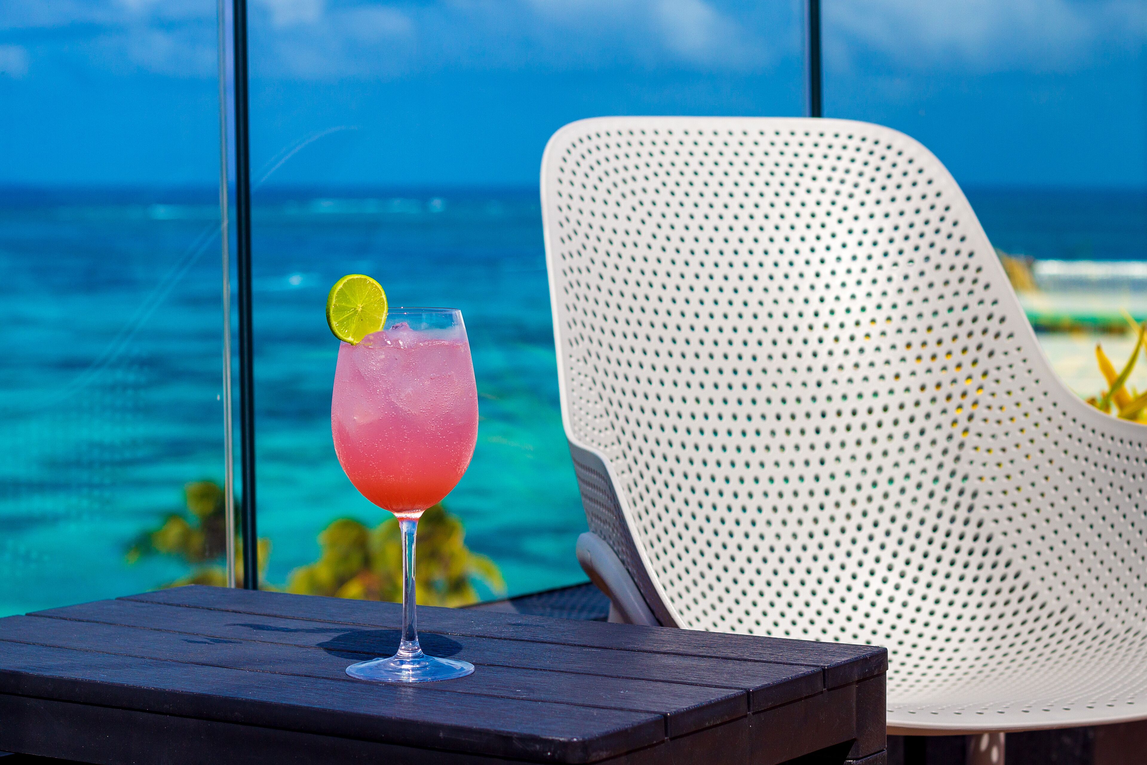 A glass of pink drink with a lemon slice on a wooden table with a view of the ocean.
