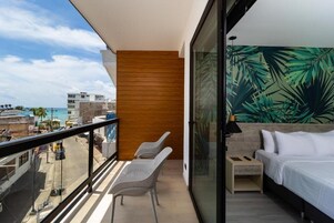 Un balcón con vista a la ciudad, una cama con ropa blanca y una pared con estampado de hojas tropicales.