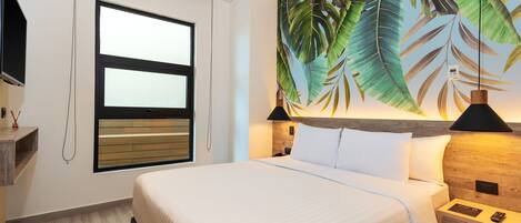 A hotel room with a large bed, a window with blinds, a wall-mounted TV, a nightstand with a lamp, and a tropical leaf mural.