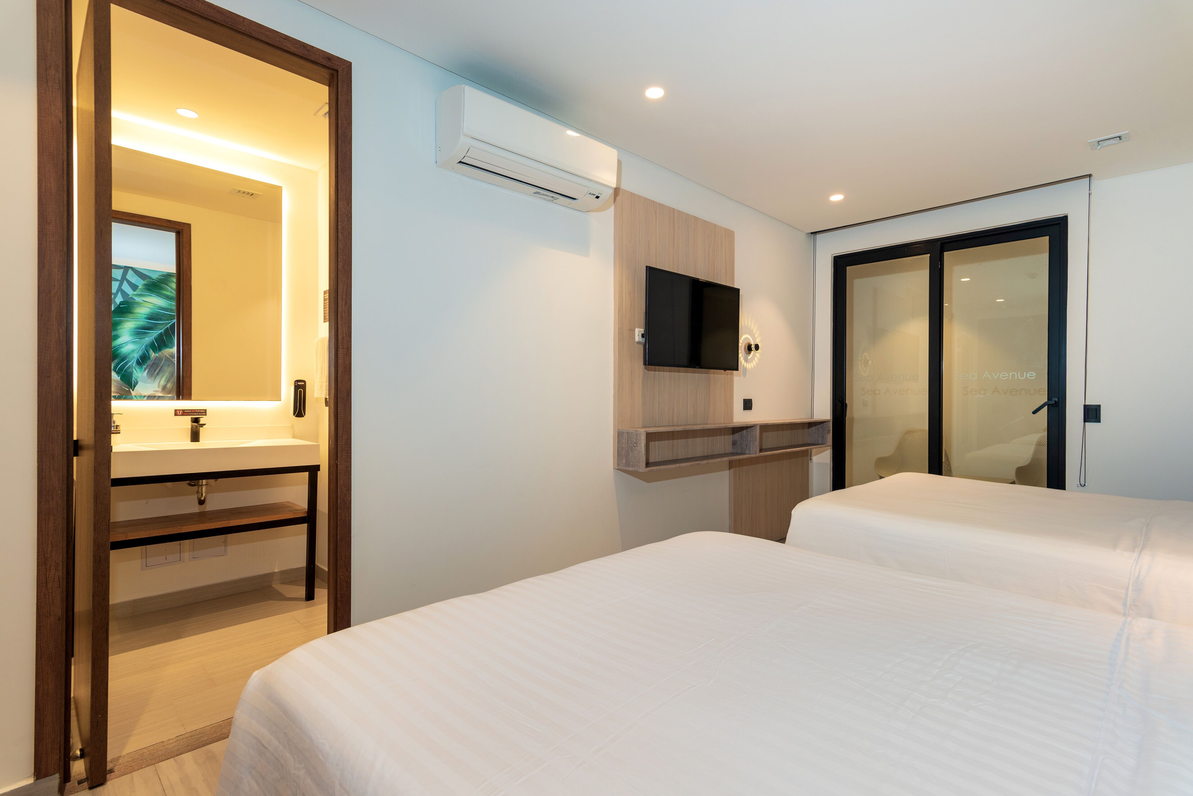 Quarto de hotel com porta de madeira que dá para um banheiro, uma TV montada na parede e uma cama com roupa de cama branca.
