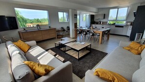 Un salon moderne comprenant un canapé, une table basse et un coin repas. De grandes fenêtres laissent entrer la lumière naturelle et offrent une vue sur la verdure environnante.
