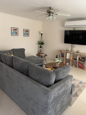 A living room with a grey sectional sofa, a ceiling fan, a flat-screen TV, a floor lamp, and a bookshelf. - Beautiful gated spacious condo 5 minute walk to beach, restaurants and Hip Strip (Montego Bay)