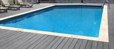 A rectangular swimming pool with a wooden deck, surrounded by a white fence, and a building with a red-tiled roof in the background.