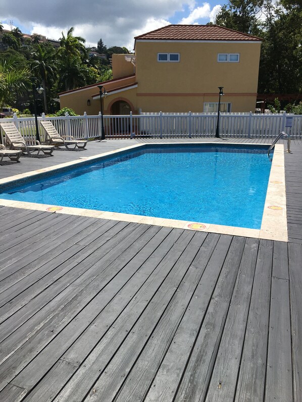 A rectangular swimming pool with a wooden deck, surrounded by a white fence, and a building with a red-tiled roof in the background. - Beautiful gated spacious condo 5 minute walk to beach, restaurants and Hip Strip (Montego Bay)