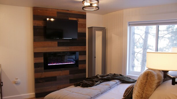 A bedroom with a fireplace, a TV mounted on a wooden wall, a tall cabinet, and a window with a view of trees. - Cozy and Clean Park City Studio (Park City)