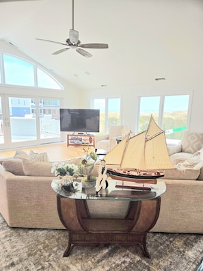 A living room with a large window, a ceiling fan, a flat-screen TV, a model sailboat, and a sofa. - Renovated Luxury Home w/Ocean & Bay Views in Gated Community (Fenwick Island)