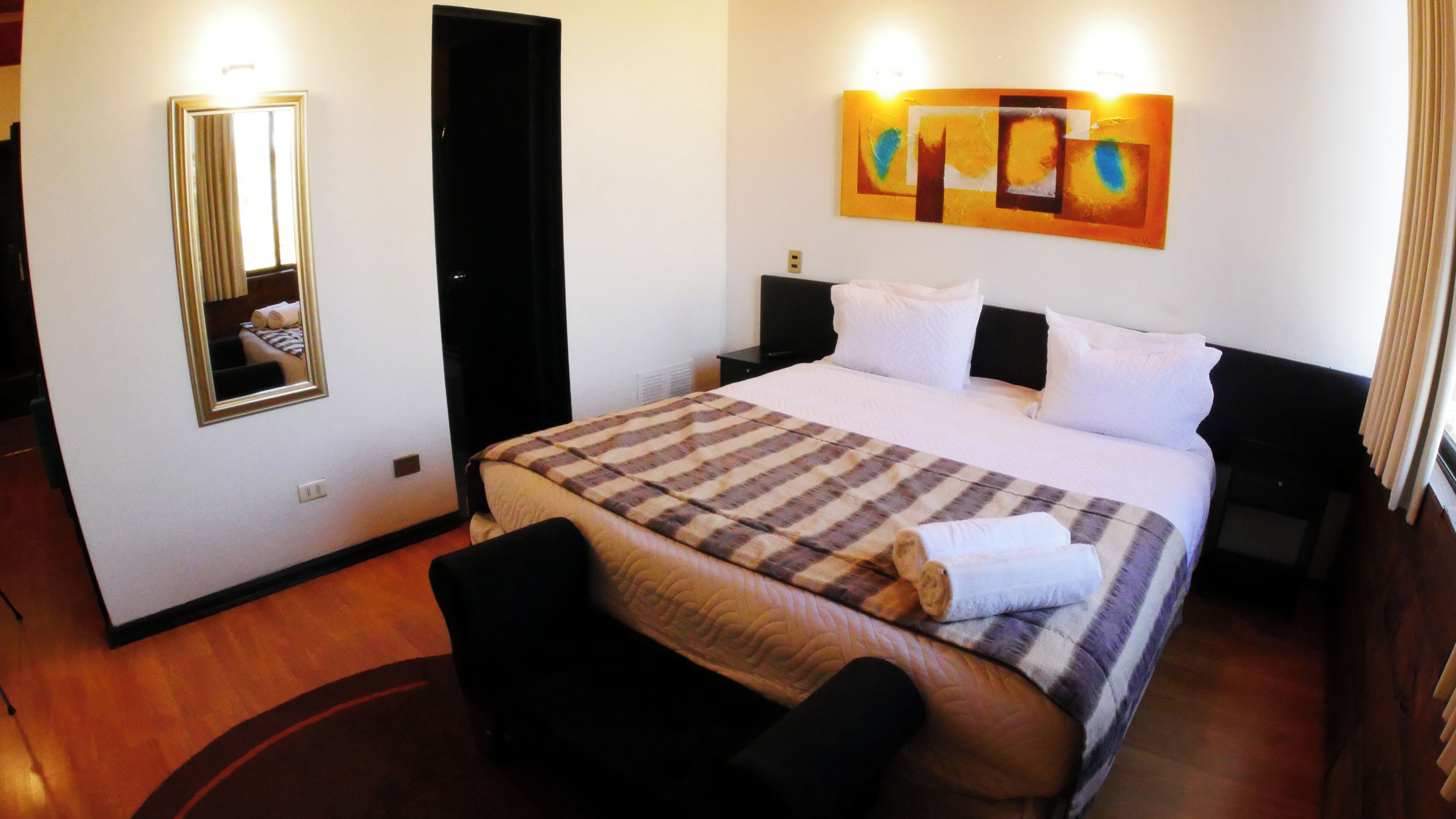 A hotel room with a bed, a chair, a nightstand, a bedside table, a mirror, and a wall art piece.