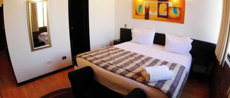 A hotel room with a bed, a chair, a nightstand, a bedside table, a mirror, and a wall art piece.