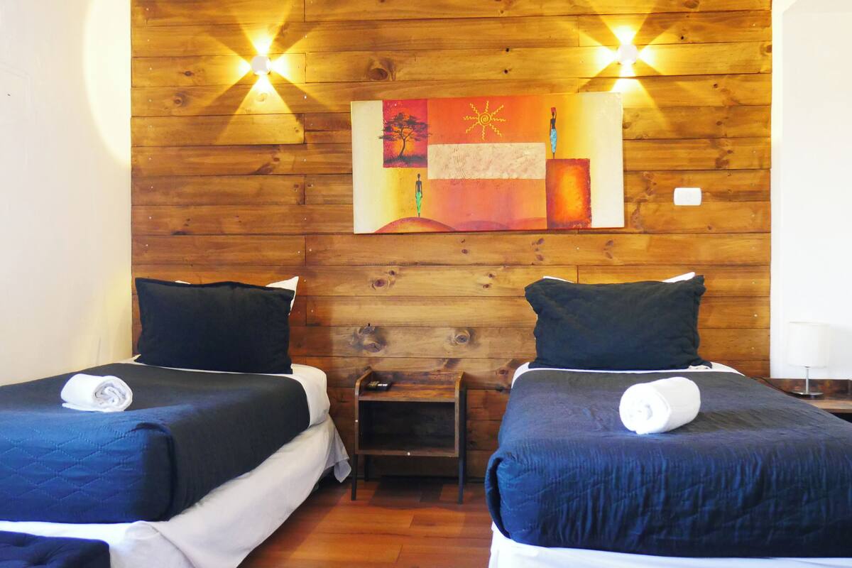 A room with two beds, wooden walls, a painting, and a small table.