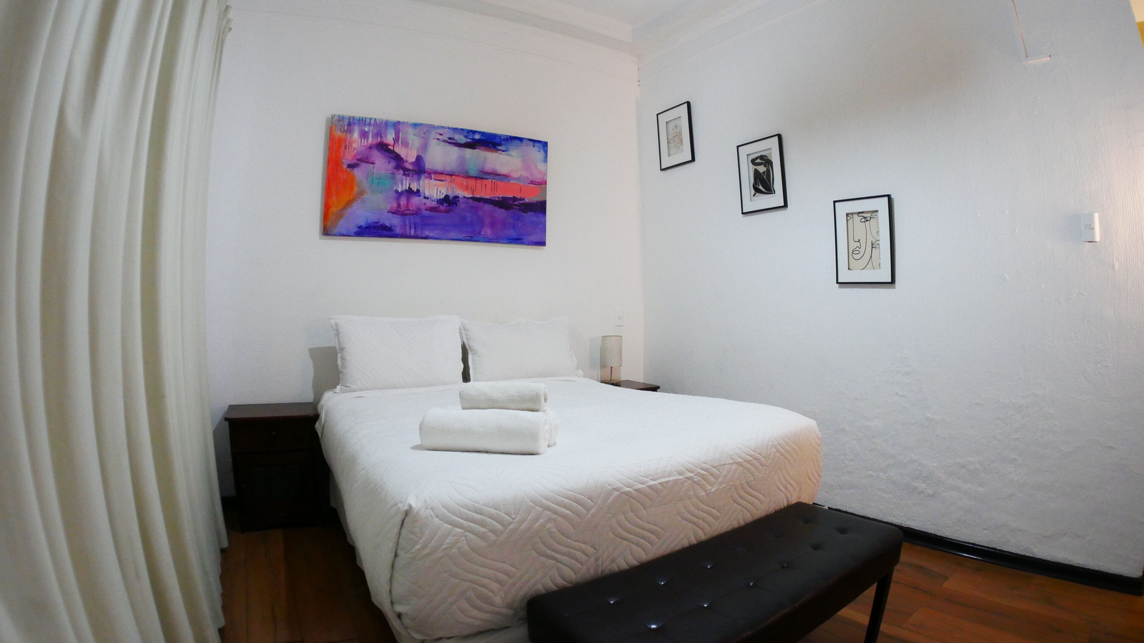A hotel room with a bed, a bench, a painting, and framed artwork on the wall.