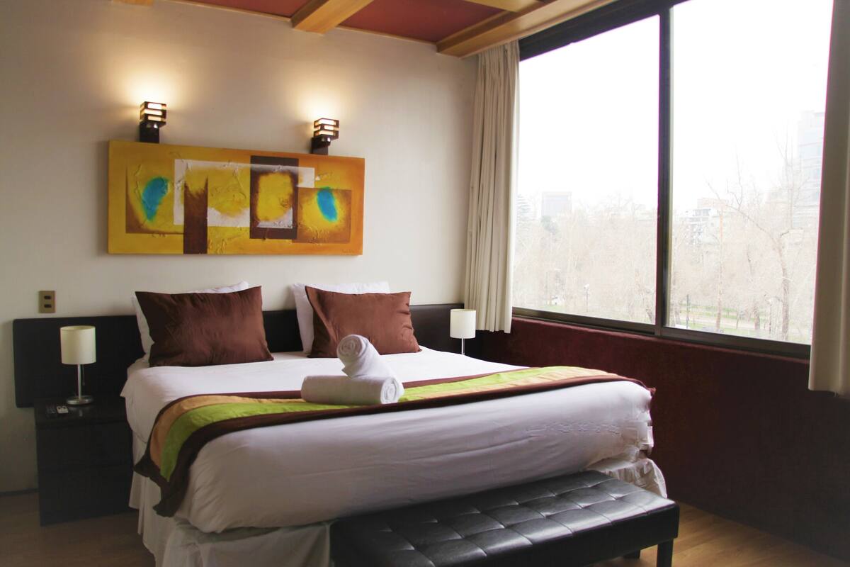 A hotel room with a bed, a bench, a nightstand, a lamp, and a painting on the wall.