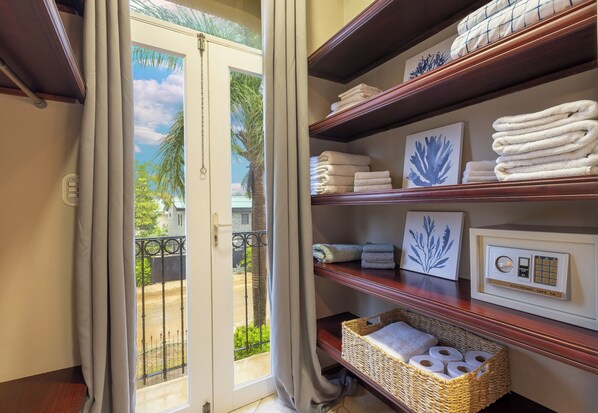 A hotel room with a balcony, a door with curtains, shelves with towels and a safe, and a basket with toiletries.