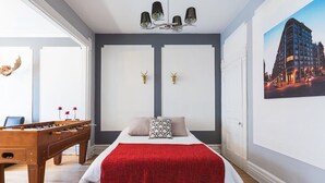 Un dormitorio con cama, una colcha roja, una mesa de fútbol de madera y un cuadro de un paisaje urbano en la pared.