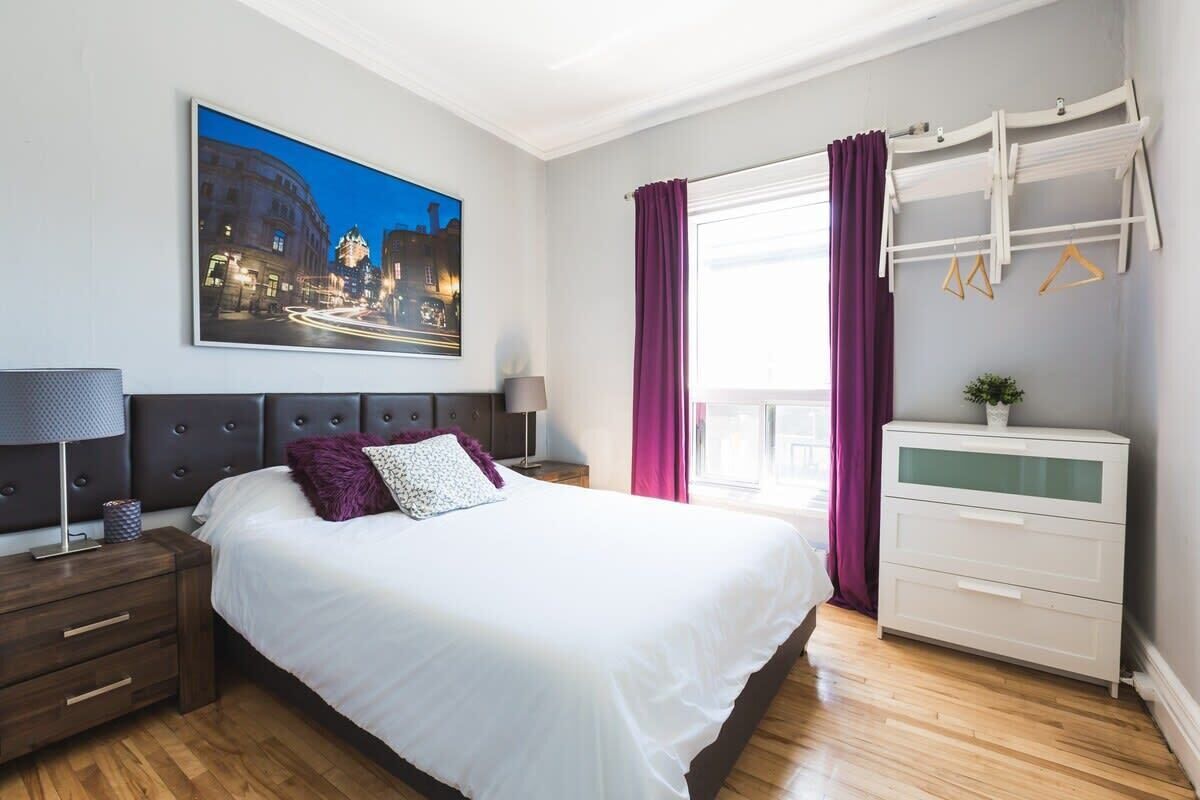 A modern bedroom with a large bed, a nightstand, a dresser, a window with curtains, and a cityscape painting on the wall.