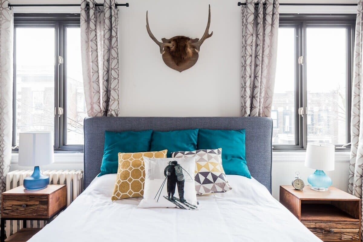 Une chambre avec un grand lit, de nombreux oreillers, une tête d’animal empaillée accrochée au mur et deux fenêtres avec des rideaux.