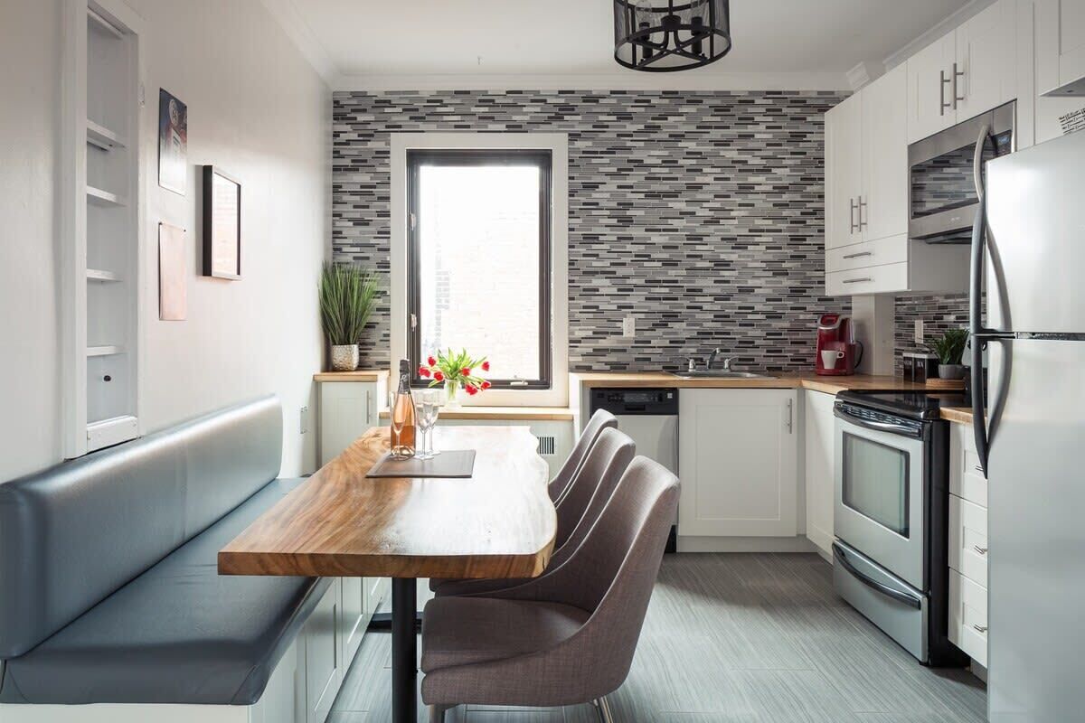 A modern kitchen with a wooden dining table, grey upholstered bench, white cabinets, and a patterned backsplash.