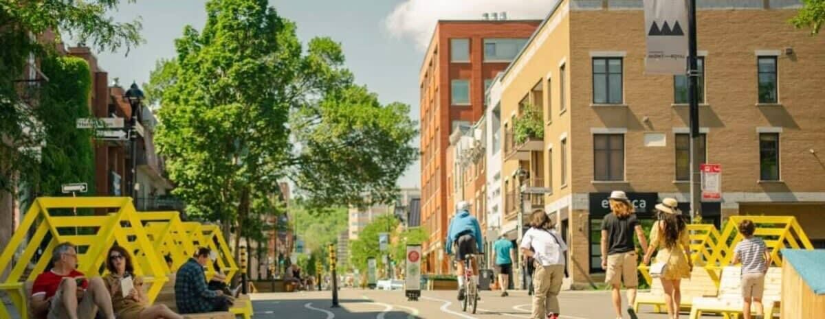Escena urbana con gente caminando y andando en bicicleta, edificios con balcones y una estructura amarilla en la acera.
