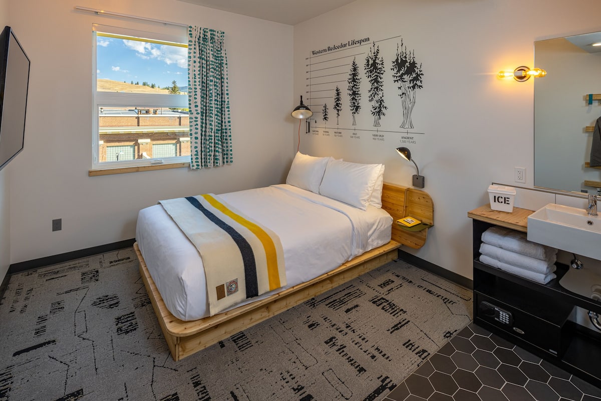 A bedroom with a bed, a window with curtains, a wall chart, a bedside lamp, a wooden nightstand, and a bathroom with a sink and towel rack.