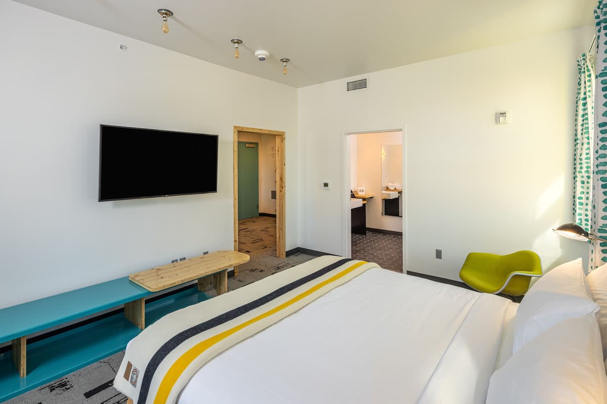 A hotel room with a bed, a TV mounted on the wall, a wooden bench, a small table, and a bright yellow chair.