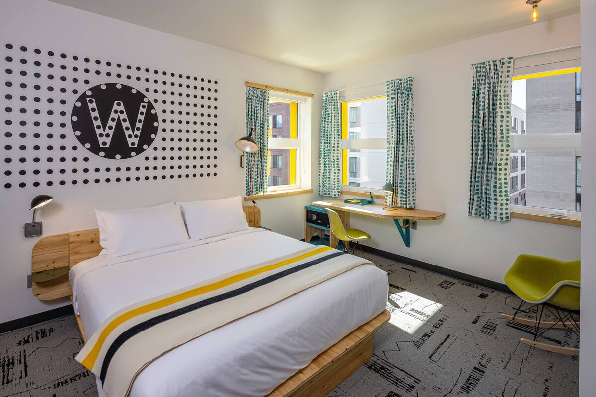 A modern hotel room with a large bed, a desk, a chair, and a wall with a large letter 'W'.