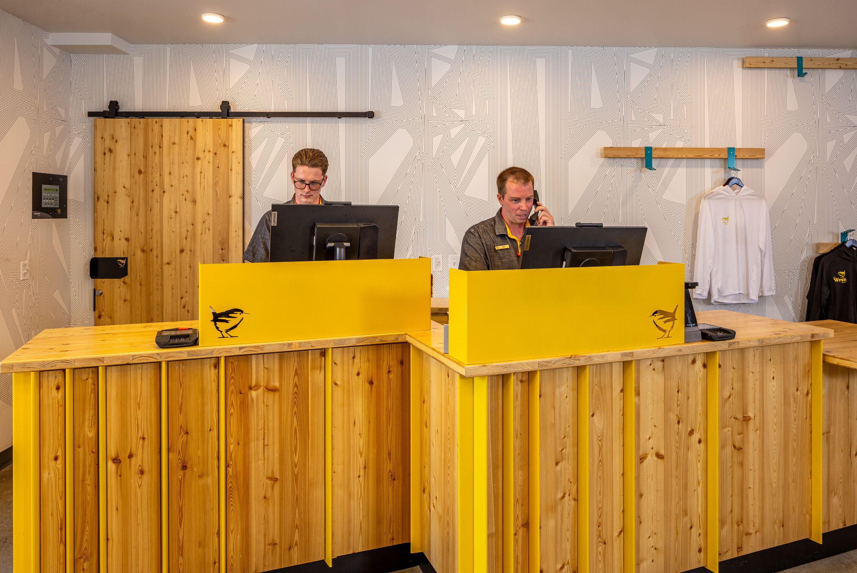 A reception desk with wooden panels and a yellow accent, two individuals at computers, and a patterned wall in the background.