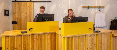 A reception desk with wooden panels and a yellow accent, two individuals at computers, and a patterned wall in the background.