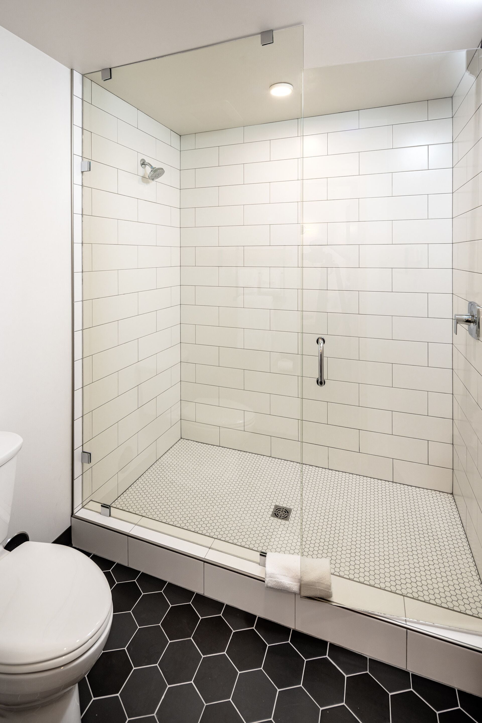 Casa de banho moderna com cabine de duche envidraçada, paredes revestidas a mosaico branco e chão com padrão hexagonal em preto e branco.