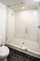 Casa de banho moderna com cabine de duche envidraçada, paredes revestidas a mosaico branco e chão com padrão hexagonal em preto e branco.