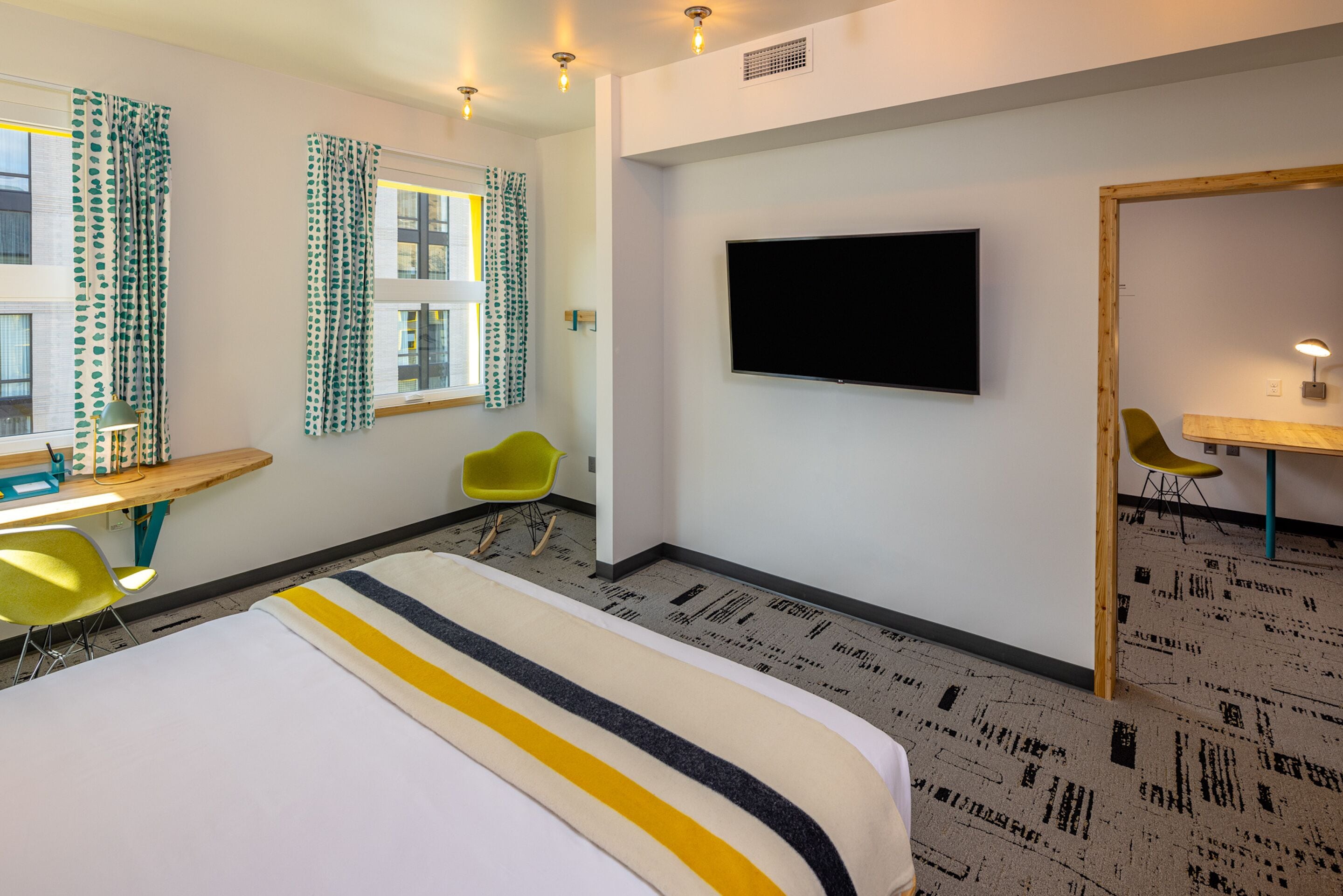 A hotel room with a bed, a desk, two chairs, a television, and a patterned carpet.