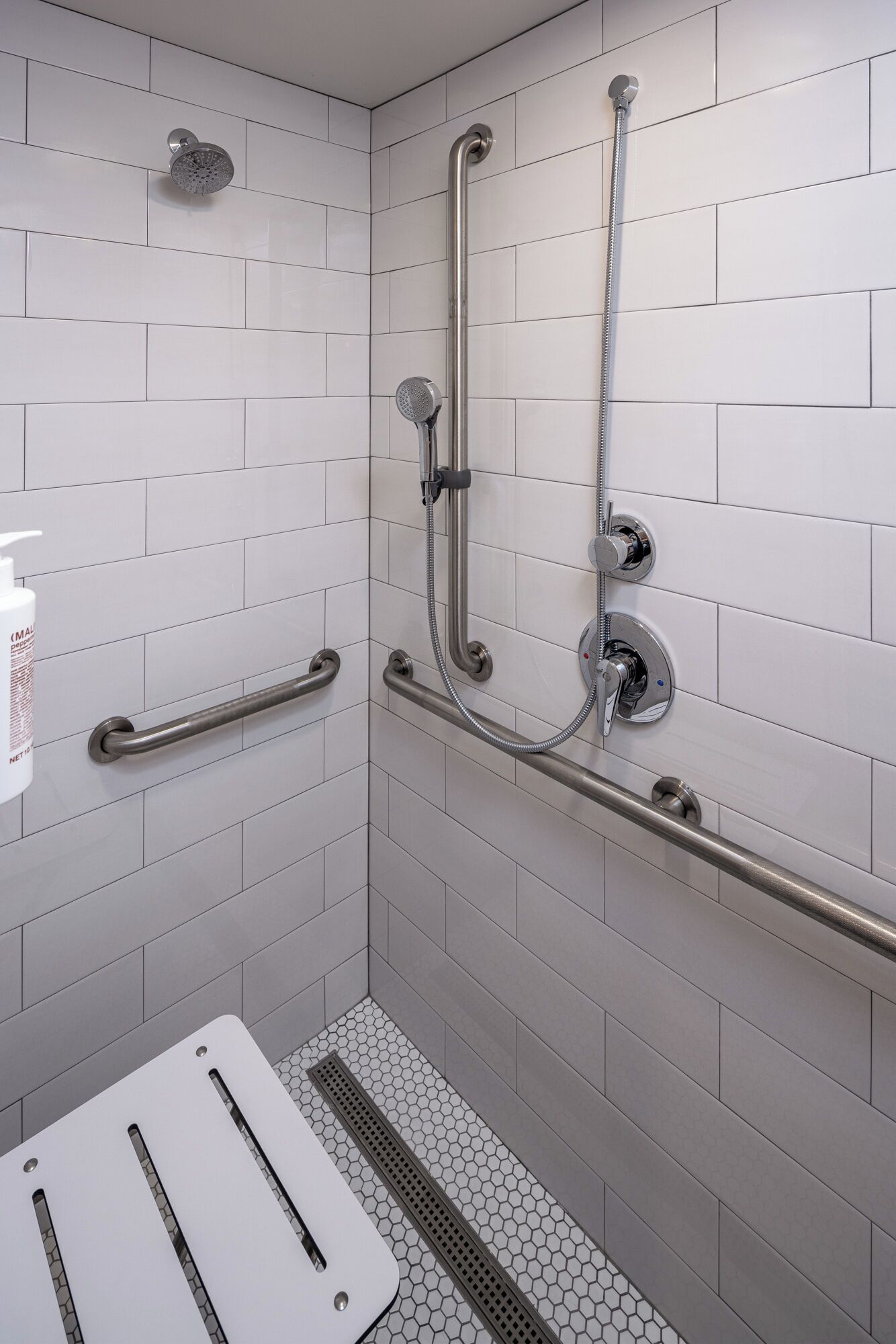 A modern bathroom with white tiled walls, a shower with a handheld showerhead, grab bars, and a white sink with a soap dispenser.
