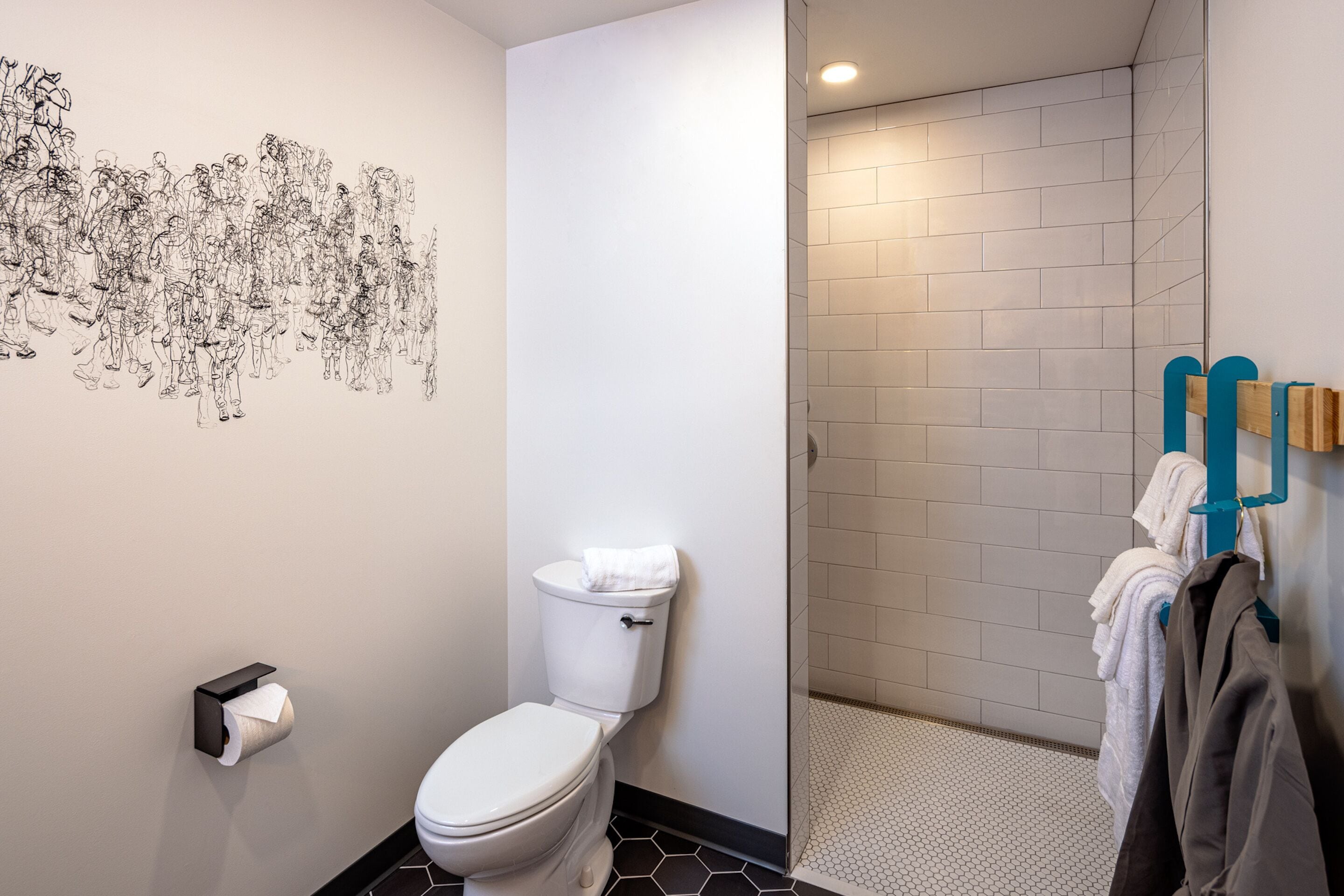 Une salle de bain équipée de toilettes, d’un sèche-mains, d’un porte-serviettes avec des serviettes, et d’un espace douche dont les murs et le sol sont recouverts de carreaux.