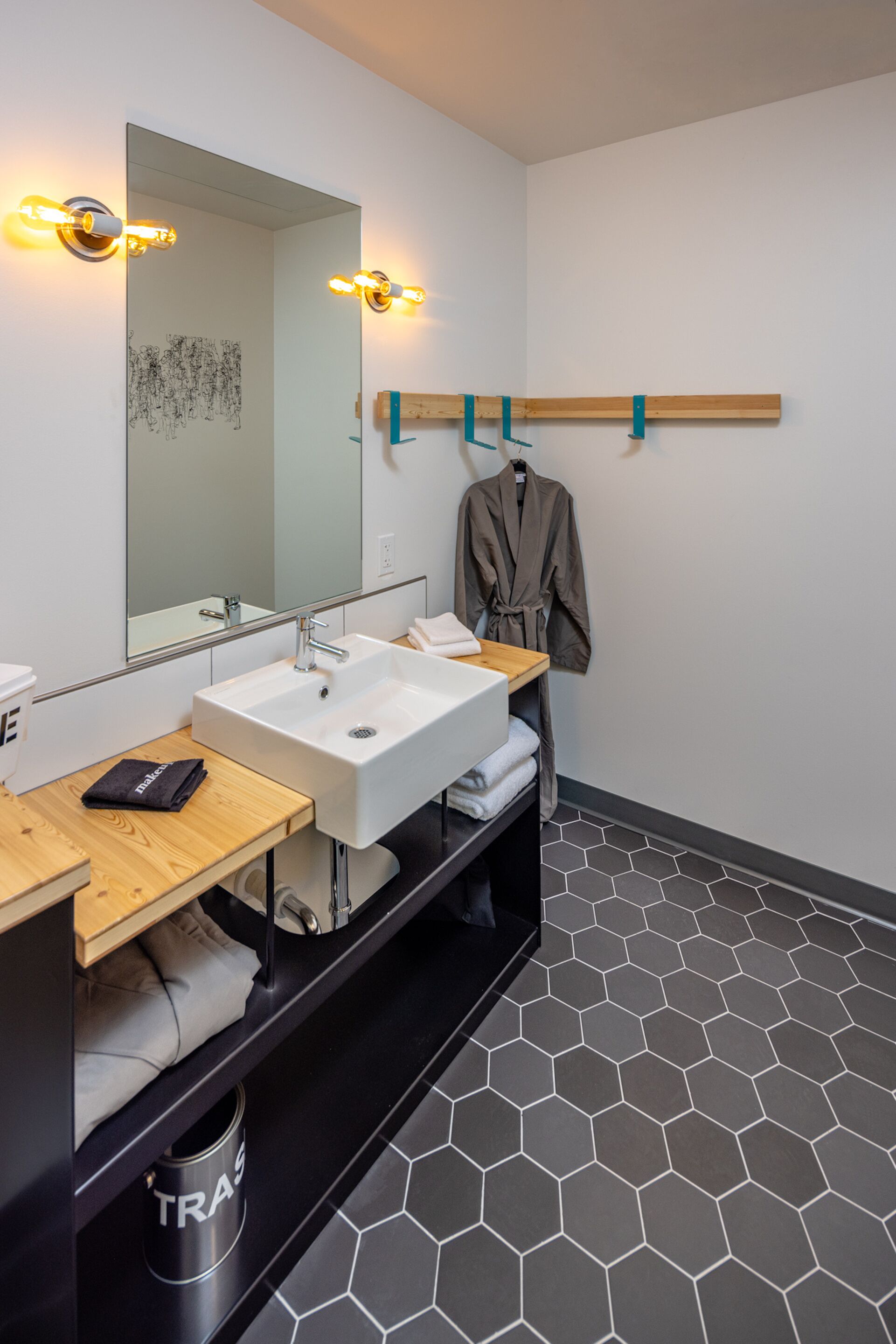 A modern bathroom with a large mirror, two sinks, and a wooden shelf with towels and a robe.