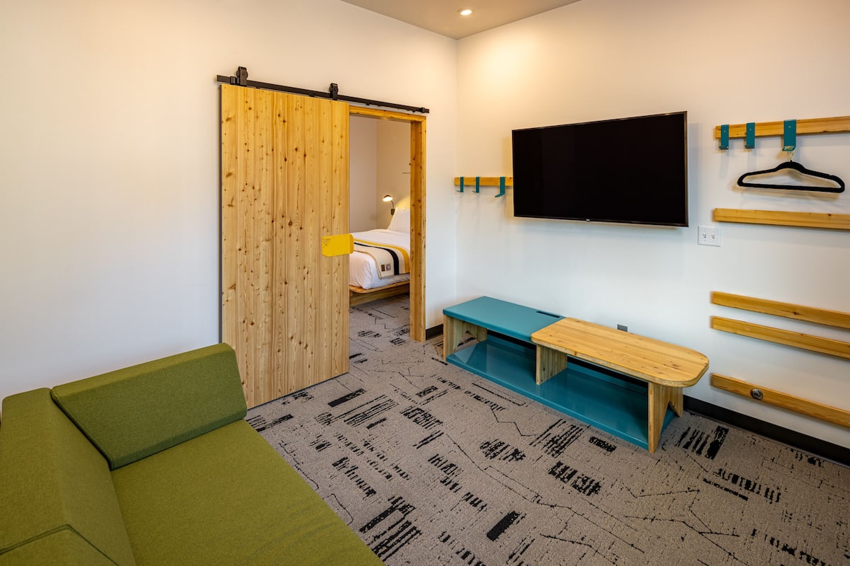 A modern room with a green sofa, a wooden bench, a TV mounted on the wall, and a sliding door leading to a bed.