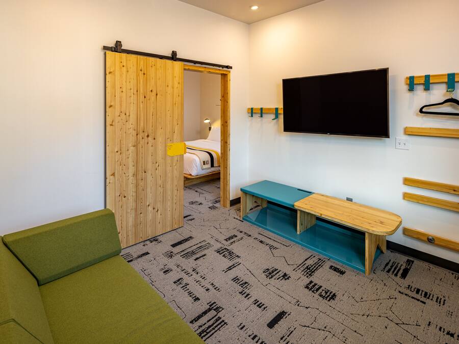 Quarto moderno com sofá verde, um banco de madeira, TV fixada na parede e porta deslizante que dá para uma cama.
