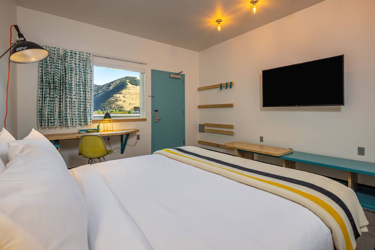 A hotel room with a bed, desk, chair, TV, and a window with a view of the mountains.