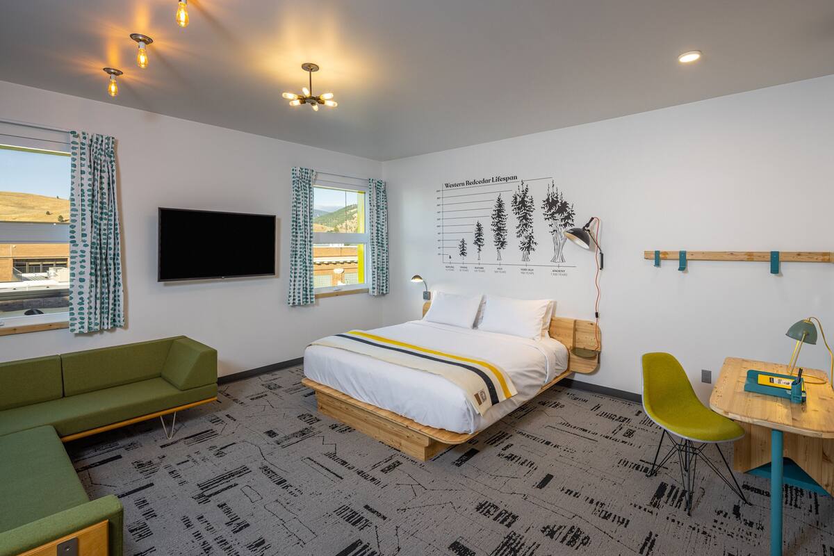 A modern hotel room with a bed, green sofa, wooden desk, and a large window with curtains.