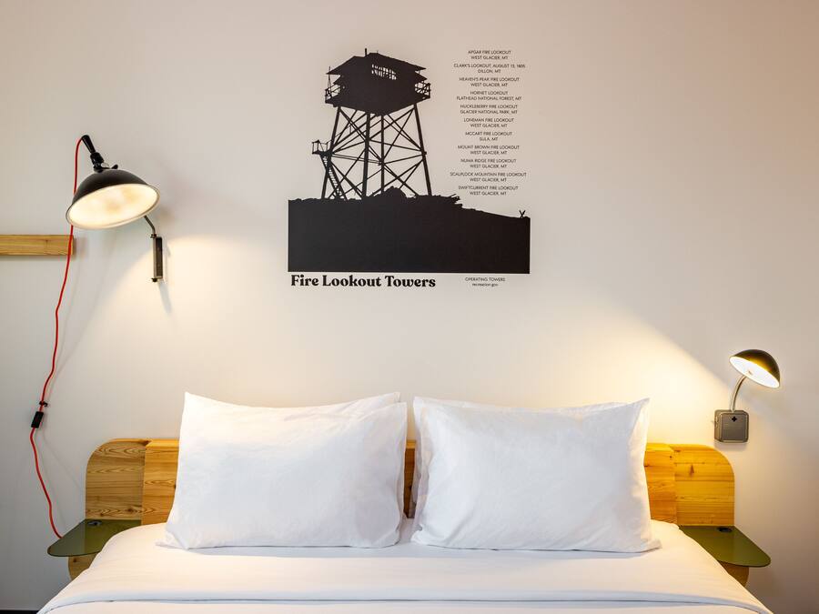 A bed with white bedding, a wall-mounted lamp, and a poster of fire lookout towers.