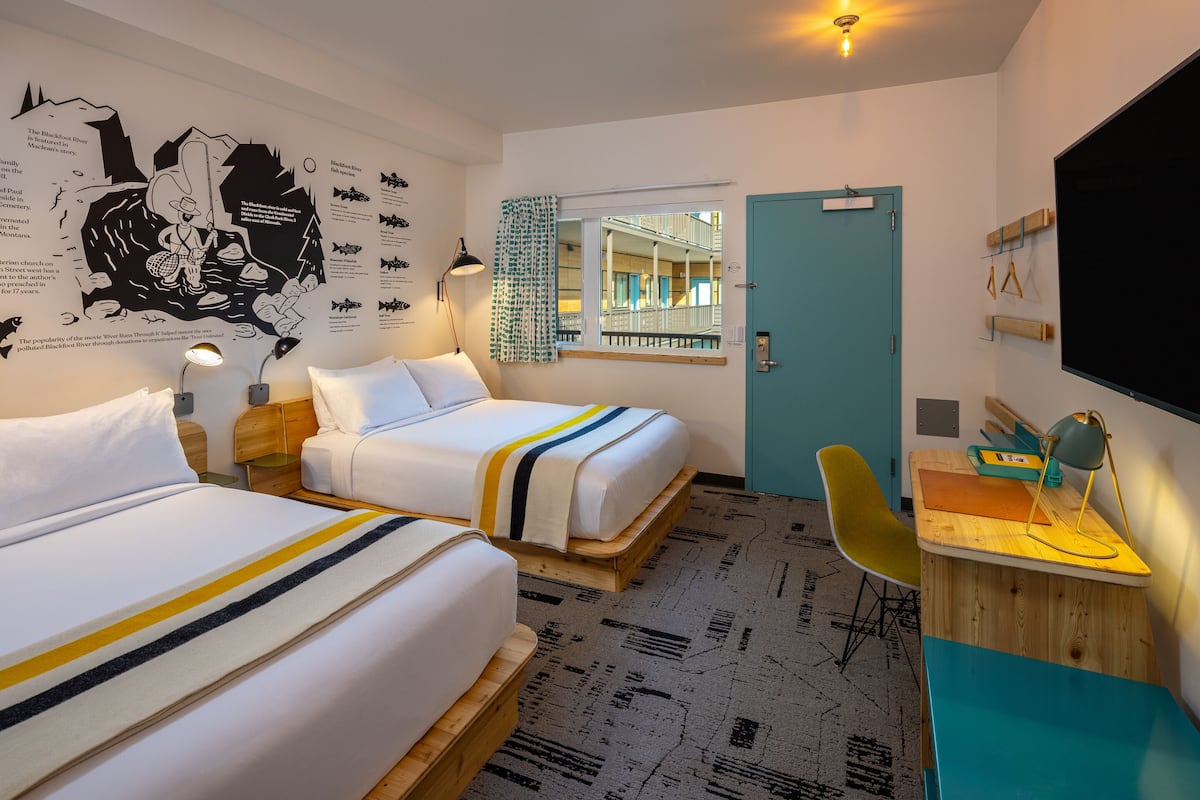 A hotel room with two beds, a desk with a chair, a TV, a window with a view of the building, and a wall with a map and text.