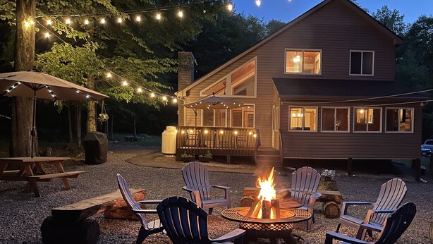 Un área de estar acogedora al aire libre con sillas Adirondack y un brasero, rodeada por una terraza de madera y luces colgantes.