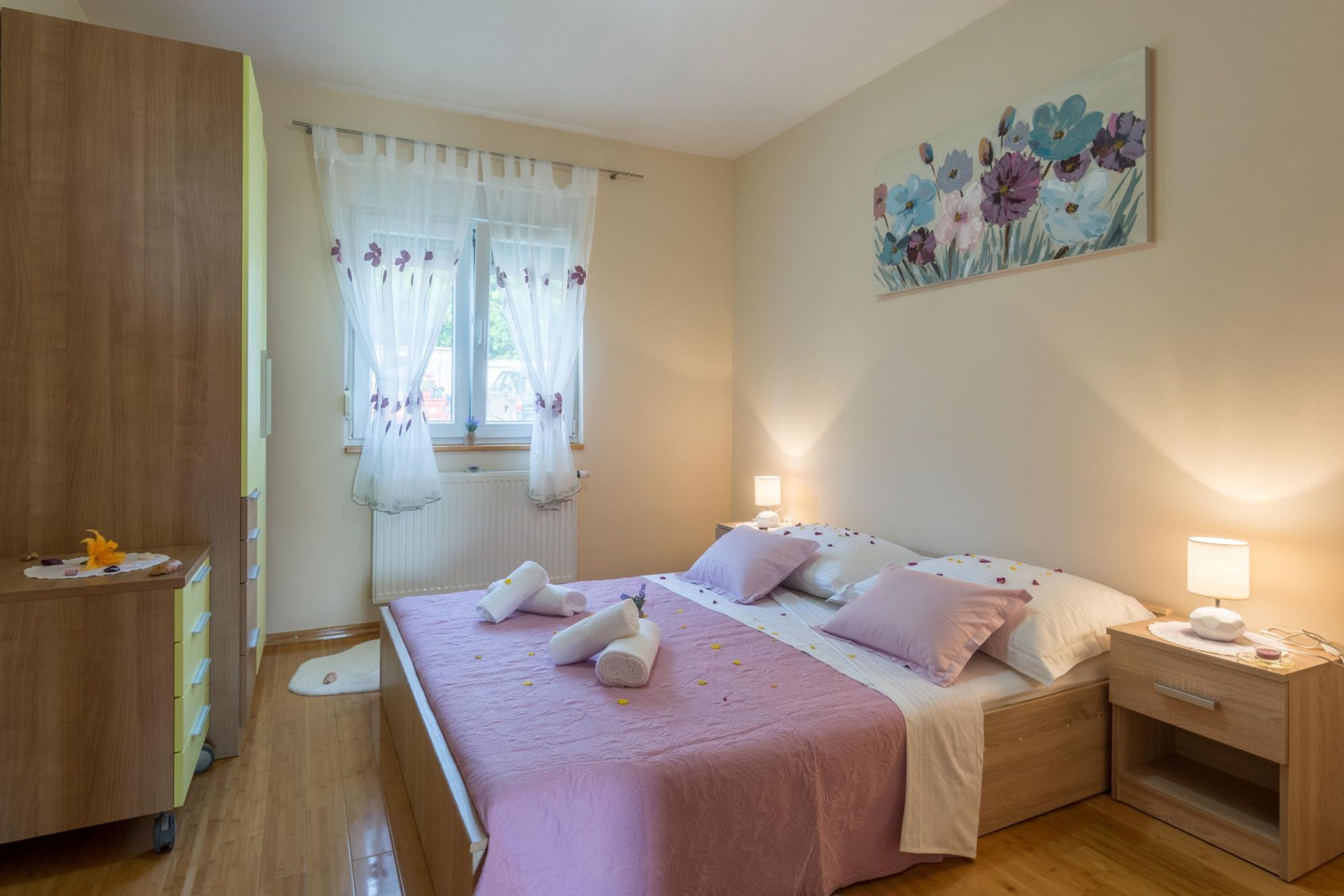 Un dormitorio con cama, mesita de noche, cómoda de madera, ventana con cortinas translúcidas y un cuadro floral en la pared.