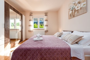 A bedroom with a large bed, a window with curtains, a mirror, and a painting on the wall.